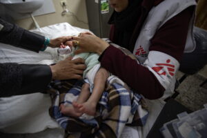 Bébé nouveau-né couché sur une table d’examen à Gaza, enveloppé dans une couverture à carreaux marron et bleu. Une soignante portant un gilet blanc et bordeaux marqué du logo Médecins Sans Frontières tient délicatement l’enfant par le torse et les épaules pour ajuster ses vêtements. On voit également les mains d’un autre adulte qui soutiennent doucement la tête du bébé. À l’arrière-plan, un évier fixé au mur et, à droite, une trousse médicale posée sur un meuble.