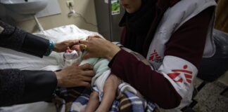 Bébé nouveau-né couché sur une table d’examen à Gaza, enveloppé dans une couverture à carreaux marron et bleu. Une soignante portant un gilet blanc et bordeaux marqué du logo Médecins Sans Frontières tient délicatement l’enfant par le torse et les épaules pour ajuster ses vêtements. On voit également les mains d’un autre adulte qui soutiennent doucement la tête du bébé. À l’arrière-plan, un évier fixé au mur et, à droite, une trousse médicale posée sur un meuble.
