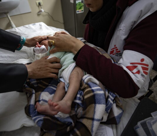 Gaza : ces organisations humanitaires sont toujours actives Bébé nouveau-né couché sur une table d’examen à Gaza, enveloppé dans une couverture à carreaux marron et bleu. Une soignante portant un gilet blanc et bordeaux marqué du logo Médecins Sans Frontières tient délicatement l’enfant par le torse et les épaules pour ajuster ses vêtements. On voit également les mains d’un autre adulte qui soutiennent doucement la tête du bébé. À l’arrière-plan, un évier fixé au mur et, à droite, une trousse médicale posée sur un meuble.