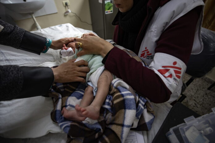 Bébé nouveau-né couché sur une table d’examen à Gaza, enveloppé dans une couverture à carreaux marron et bleu. Une soignante portant un gilet blanc et bordeaux marqué du logo Médecins Sans Frontières tient délicatement l’enfant par le torse et les épaules pour ajuster ses vêtements. On voit également les mains d’un autre adulte qui soutiennent doucement la tête du bébé. À l’arrière-plan, un évier fixé au mur et, à droite, une trousse médicale posée sur un meuble.