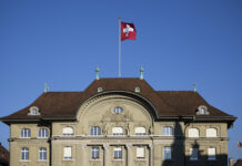 « Les bénéfices de la Banque nationale appartiennent au peuple » Façade de la Banque nationale suisse à Berne, bâtiment classique à toiture en tuiles brunes, drapeau suisse flottant sur un mât central, ciel bleu en arrière-plan.