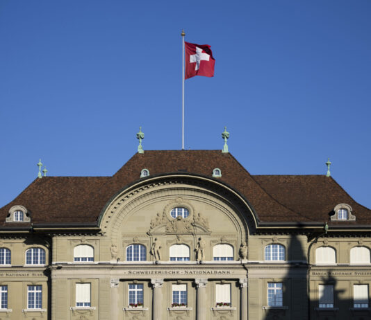 « Les bénéfices de la Banque nationale appartiennent au peuple » Façade de la Banque nationale suisse à Berne, bâtiment classique à toiture en tuiles brunes, drapeau suisse flottant sur un mât central, ciel bleu en arrière-plan.