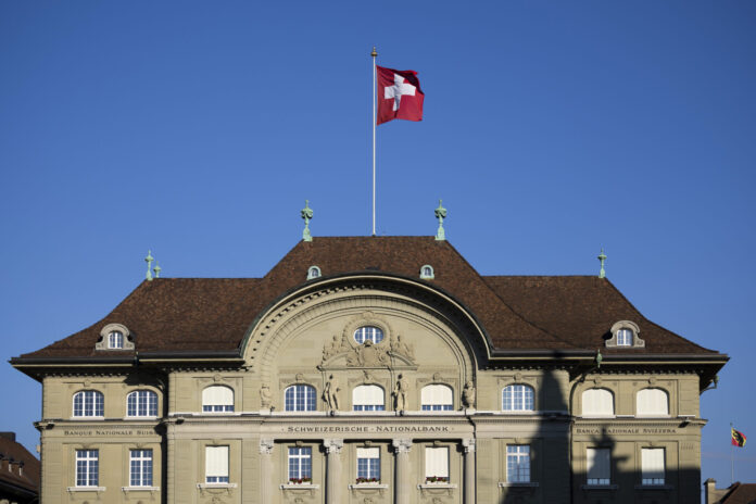Façade de la Banque nationale suisse à Berne, bâtiment classique à toiture en tuiles brunes, drapeau suisse flottant sur un mât central, ciel bleu en arrière-plan.