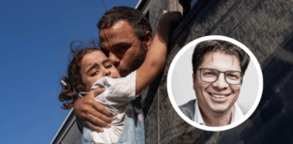 Sous un ciel bleu, un homme penché par la fenêtre d’un autocar sombre serre une petite fille dans ses bras et l’embrasse sur la joue. La fillette, vêtue d’une salopette en jean et d’un t-shirt blanc, agrippe le rebord du véhicule, le regard mélancolique, tandis qu’un autre passager observe la scène en arrière-plan.