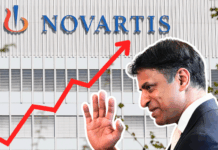 Médicaments : Novartis demande encore plus d’argent malgré des prix déjà élevés Montage photographique : en arrière-plan, la façade d’un immeuble gris clair arbore en grand le logo et le nom « NOVARTIS ». En surimpression, une flèche rouge zigzaguant de l’angle inférieur gauche vers l’angle supérieur droit symbolise une forte progression. Au premier plan, sur la droite, un homme en costume sombre, de profil, lève la main comme pour saluer.