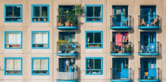 Façade d’un immeuble résidentiel aux lignes géométriques marquées : trois étages de larges fenêtres carrées encadrées de bleu vif, alternant avec des balcons aux rambardes noires. Chaque balcon dévoile un petit espace de vie extérieur : chaises pliantes, tables d’appoint, plantes en pots, quelques vêtements séchant sur un fil, parfois un fauteuil ou un vieux dossier de canapé. Derrière les fenêtres, on devine rideaux, bibliothèques et lampes d’intérieur. Le sol est bordé d’une mince bande de trottoir où poussent quelques brins d’herbe et de petites fleurs sauvages.