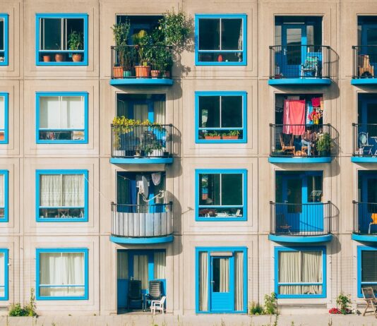 Quatre leviers pour réduire la pression sur les locataires : entre régulation, logements sociaux et contrôle des loyers Façade d’un immeuble résidentiel aux lignes géométriques marquées : trois étages de larges fenêtres carrées encadrées de bleu vif, alternant avec des balcons aux rambardes noires. Chaque balcon dévoile un petit espace de vie extérieur : chaises pliantes, tables d’appoint, plantes en pots, quelques vêtements séchant sur un fil, parfois un fauteuil ou un vieux dossier de canapé. Derrière les fenêtres, on devine rideaux, bibliothèques et lampes d’intérieur. Le sol est bordé d’une mince bande de trottoir où poussent quelques brins d’herbe et de petites fleurs sauvages.