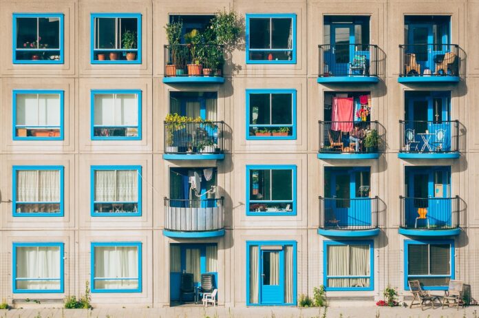 Façade d’un immeuble résidentiel aux lignes géométriques marquées : trois étages de larges fenêtres carrées encadrées de bleu vif, alternant avec des balcons aux rambardes noires. Chaque balcon dévoile un petit espace de vie extérieur : chaises pliantes, tables d’appoint, plantes en pots, quelques vêtements séchant sur un fil, parfois un fauteuil ou un vieux dossier de canapé. Derrière les fenêtres, on devine rideaux, bibliothèques et lampes d’intérieur. Le sol est bordé d’une mince bande de trottoir où poussent quelques brins d’herbe et de petites fleurs sauvages.