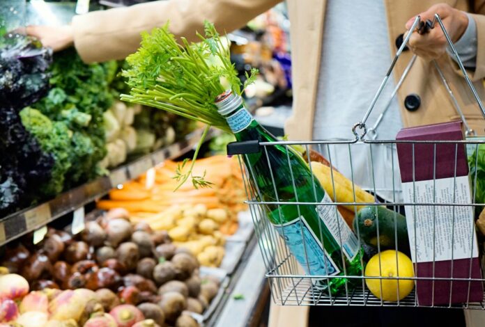 Dans une épicerie, une personne vêtue d’un manteau beige tient un panier métallique rempli d’une bouteille d’eau gazeuse, d’un bouquet de fanes de carottes, d’un citron, d’une courgette et de quelques produits emballés. Elle se tient devant les étals garnis de choux, de brocolis, de betteraves, de navets et de carottes.