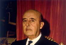 Cinquante ans après Franco : quand les crises ravivent le danger fasciste Portrait de Franco, chauve, au visage sérieux, vêtu d'un costume sombre avec une chemise blanche et une cravate noire. Il est assis sur un fauteuil orné de motifs dorés, devant un rideau rouge foncé.