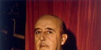 Cinquante ans après Franco : quand les crises ravivent le danger fasciste Portrait de Franco, chauve, au visage sérieux, vêtu d'un costume sombre avec une chemise blanche et une cravate noire. Il est assis sur un fauteuil orné de motifs dorés, devant un rideau rouge foncé.