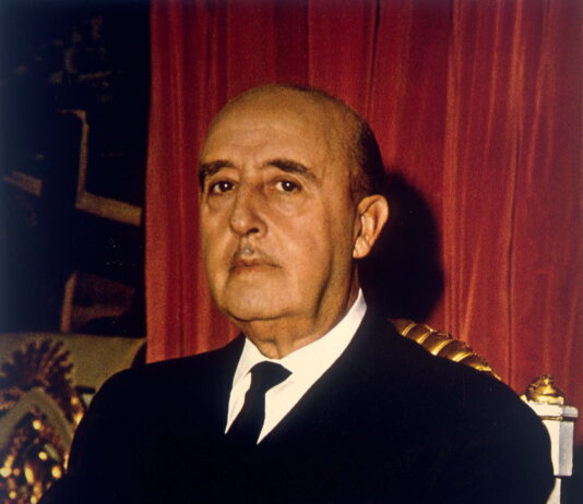 Cinquante ans après Franco : quand les crises ravivent le danger fasciste Portrait de Franco, chauve, au visage sérieux, vêtu d'un costume sombre avec une chemise blanche et une cravate noire. Il est assis sur un fauteuil orné de motifs dorés, devant un rideau rouge foncé.