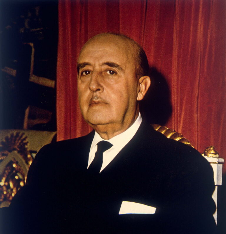 Portrait de Franco, chauve, au visage sérieux, vêtu d'un costume sombre avec une chemise blanche et une cravate noire. Il est assis sur un fauteuil orné de motifs dorés, devant un rideau rouge foncé.