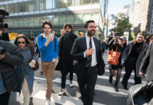 Zohran Mamdani est le nouveau maire de la ville de New York Sur un large passage piéton en pleine ville de New York, Zohran Mamdani en costume sombre, chemise blanche et cravate marche au centre du groupe, un grand sourire aux lèvres et la main posée sur la poitrine. Autour de lui, plusieurs personnes le suivent de près : à gauche, un caméraman professionnel filme avec une grosse caméra et une femme en trench-coat bleu et lunettes de soleil avance en tenant son téléphone ; à droite, deux jeunes femmes souriantes, l’une casquette vissée sur la tête et un grand sac orange sur l’épaule, l’autre dans un manteau à carreaux, filment ou photographient la scène avec leur smartphone. En arrière-plan, on devine des façades vitrées d’immeubles de bureaux et quelques passants sur le trottoir.