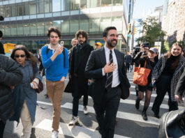 Zohran Mamdani est le nouveau maire de la ville de New York Sur un large passage piéton en pleine ville de New York, Zohran Mamdani en costume sombre, chemise blanche et cravate marche au centre du groupe, un grand sourire aux lèvres et la main posée sur la poitrine. Autour de lui, plusieurs personnes le suivent de près : à gauche, un caméraman professionnel filme avec une grosse caméra et une femme en trench-coat bleu et lunettes de soleil avance en tenant son téléphone ; à droite, deux jeunes femmes souriantes, l’une casquette vissée sur la tête et un grand sac orange sur l’épaule, l’autre dans un manteau à carreaux, filment ou photographient la scène avec leur smartphone. En arrière-plan, on devine des façades vitrées d’immeubles de bureaux et quelques passants sur le trottoir.