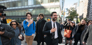 Zohran Mamdani est le nouveau maire de la ville de New York Sur un large passage piéton en pleine ville de New York, Zohran Mamdani en costume sombre, chemise blanche et cravate marche au centre du groupe, un grand sourire aux lèvres et la main posée sur la poitrine. Autour de lui, plusieurs personnes le suivent de près : à gauche, un caméraman professionnel filme avec une grosse caméra et une femme en trench-coat bleu et lunettes de soleil avance en tenant son téléphone ; à droite, deux jeunes femmes souriantes, l’une casquette vissée sur la tête et un grand sac orange sur l’épaule, l’autre dans un manteau à carreaux, filment ou photographient la scène avec leur smartphone. En arrière-plan, on devine des façades vitrées d’immeubles de bureaux et quelques passants sur le trottoir.