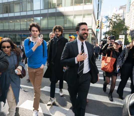 Zohran Mamdani est le nouveau maire de la ville de New York Sur un large passage piéton en pleine ville de New York, Zohran Mamdani en costume sombre, chemise blanche et cravate marche au centre du groupe, un grand sourire aux lèvres et la main posée sur la poitrine. Autour de lui, plusieurs personnes le suivent de près : à gauche, un caméraman professionnel filme avec une grosse caméra et une femme en trench-coat bleu et lunettes de soleil avance en tenant son téléphone ; à droite, deux jeunes femmes souriantes, l’une casquette vissée sur la tête et un grand sac orange sur l’épaule, l’autre dans un manteau à carreaux, filment ou photographient la scène avec leur smartphone. En arrière-plan, on devine des façades vitrées d’immeubles de bureaux et quelques passants sur le trottoir.