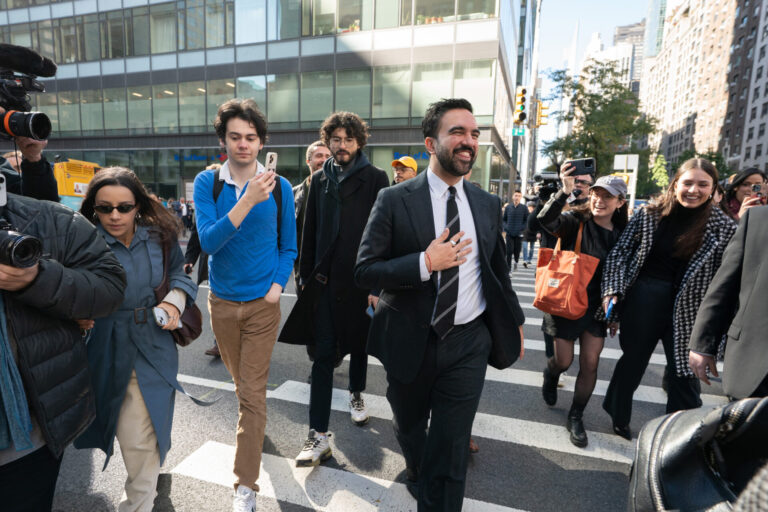 Sur un large passage piéton en pleine ville de New York, Zohran Mamdani en costume sombre, chemise blanche et cravate marche au centre du groupe, un grand sourire aux lèvres et la main posée sur la poitrine. Autour de lui, plusieurs personnes le suivent de près : à gauche, un caméraman professionnel filme avec une grosse caméra et une femme en trench-coat bleu et lunettes de soleil avance en tenant son téléphone ; à droite, deux jeunes femmes souriantes, l’une casquette vissée sur la tête et un grand sac orange sur l’épaule, l’autre dans un manteau à carreaux, filment ou photographient la scène avec leur smartphone. En arrière-plan, on devine des façades vitrées d’immeubles de bureaux et quelques passants sur le trottoir.