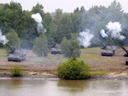 Les dépenses militaires record compromettent le développement durable Cinq canons automoteurs camouflés, alignés en position de tir sur une berge boueuse, font feu simultanément vers un plan d’eau au premier plan, projetant d’épaisses volutes de fumée blanche, avec en arrière-plan une forêt dense aux feuillages verts.