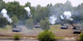 Les dépenses militaires record compromettent le développement durable Cinq canons automoteurs camouflés, alignés en position de tir sur une berge boueuse, font feu simultanément vers un plan d’eau au premier plan, projetant d’épaisses volutes de fumée blanche, avec en arrière-plan une forêt dense aux feuillages verts.