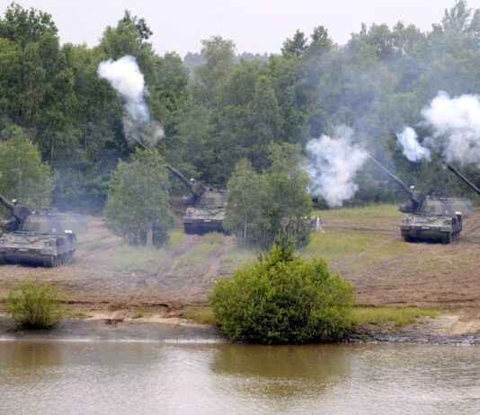 Les dépenses militaires record compromettent le développement durable Cinq canons automoteurs camouflés, alignés en position de tir sur une berge boueuse, font feu simultanément vers un plan d’eau au premier plan, projetant d’épaisses volutes de fumée blanche, avec en arrière-plan une forêt dense aux feuillages verts.