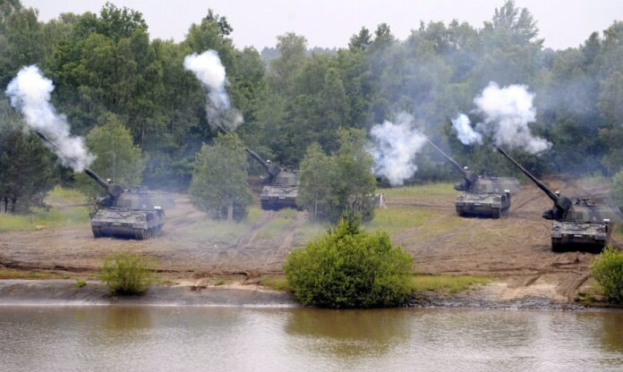 Cinq canons automoteurs camouflés, alignés en position de tir sur une berge boueuse, font feu simultanément vers un plan d’eau au premier plan, projetant d’épaisses volutes de fumée blanche, avec en arrière-plan une forêt dense aux feuillages verts.
