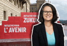 Oriane Sarrasin : « Plus on est riche, plus on pollue – c’est un fait » Portrait en buste d’Oriane Sarrasin souriante, lunettes et veste sombre, debout devant un mur de grosses boîtes rouges empilées sur le parvis d’un bâtiment en pierres clair. Sur les boîtes, on lit en blanc les inscriptions “Initiative pour l’avenir” et “Initiative für eine Zukunft”. Le ciel est nuageux et on distingue un arbre sans feuilles au loin.