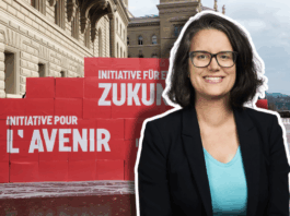 Oriane Sarrasin : « Plus on est riche, plus on pollue – c’est un fait » Portrait en buste d’Oriane Sarrasin souriante, lunettes et veste sombre, debout devant un mur de grosses boîtes rouges empilées sur le parvis d’un bâtiment en pierres clair. Sur les boîtes, on lit en blanc les inscriptions “Initiative pour l’avenir” et “Initiative für eine Zukunft”. Le ciel est nuageux et on distingue un arbre sans feuilles au loin.