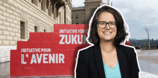 Oriane Sarrasin : « Plus on est riche, plus on pollue – c’est un fait » Portrait en buste d’Oriane Sarrasin souriante, lunettes et veste sombre, debout devant un mur de grosses boîtes rouges empilées sur le parvis d’un bâtiment en pierres clair. Sur les boîtes, on lit en blanc les inscriptions “Initiative pour l’avenir” et “Initiative für eine Zukunft”. Le ciel est nuageux et on distingue un arbre sans feuilles au loin.