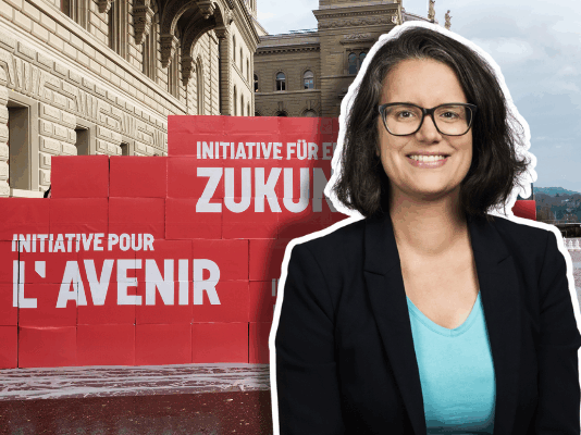 Oriane Sarrasin : « Plus on est riche, plus on pollue – c’est un fait » Portrait en buste d’Oriane Sarrasin souriante, lunettes et veste sombre, debout devant un mur de grosses boîtes rouges empilées sur le parvis d’un bâtiment en pierres clair. Sur les boîtes, on lit en blanc les inscriptions “Initiative pour l’avenir” et “Initiative für eine Zukunft”. Le ciel est nuageux et on distingue un arbre sans feuilles au loin.