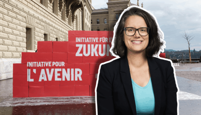 Oriane 3 (6) Portrait en buste d’Oriane Sarrasin souriante, lunettes et veste sombre, debout devant un mur de grosses boîtes rouges empilées sur le parvis d’un bâtiment en pierres clair. Sur les boîtes, on lit en blanc les inscriptions “Initiative pour l’avenir” et “Initiative für eine Zukunft”. Le ciel est nuageux et on distingue un arbre sans feuilles au loin.