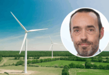 Bruno Tobback : « Le ‹ Green Deal › est également une déclaration d’indépendance » Vue aérienne d’un vaste paysage de campagne verdoyante parsemé de deux grandes éoliennes blanches sous un ciel bleu clair. En surimpression, en haut à droite, une vignette circulaire montre le visage de Bruno Tobback, un homme d’âge moyen, regardant l’objectif, coiffé court et portant une légère barbe grise.