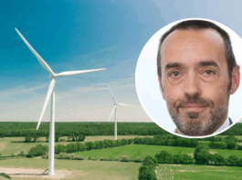 Bruno Tobback : « Le ‹ Green Deal › est également une déclaration d’indépendance » Vue aérienne d’un vaste paysage de campagne verdoyante parsemé de deux grandes éoliennes blanches sous un ciel bleu clair. En surimpression, en haut à droite, une vignette circulaire montre le visage de Bruno Tobback, un homme d’âge moyen, regardant l’objectif, coiffé court et portant une légère barbe grise.