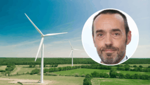 Vue aérienne d’un vaste paysage de campagne verdoyante parsemé de deux grandes éoliennes blanches sous un ciel bleu clair. En surimpression, en haut à droite, une vignette circulaire montre le visage de Bruno Tobback, un homme d’âge moyen, regardant l’objectif, coiffé court et portant une légère barbe grise.