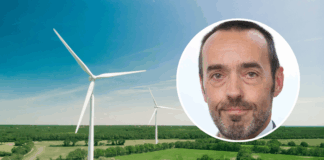 Bruno Tobback : « Le ‹ Green Deal › est également une déclaration d’indépendance » Vue aérienne d’un vaste paysage de campagne verdoyante parsemé de deux grandes éoliennes blanches sous un ciel bleu clair. En surimpression, en haut à droite, une vignette circulaire montre le visage de Bruno Tobback, un homme d’âge moyen, regardant l’objectif, coiffé court et portant une légère barbe grise.
