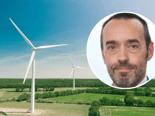 Bruno Tobback : « Le ‹ Green Deal › est également une déclaration d’indépendance » Vue aérienne d’un vaste paysage de campagne verdoyante parsemé de deux grandes éoliennes blanches sous un ciel bleu clair. En surimpression, en haut à droite, une vignette circulaire montre le visage de Bruno Tobback, un homme d’âge moyen, regardant l’objectif, coiffé court et portant une légère barbe grise.