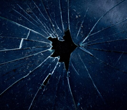 Féminicides : la réalité qui heurte Photo en gros plan d’une vitre sombre brisée, avec des fissures en éventail rayonnant à partir d’un trou central.