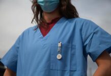Une alliance appelle à la manifestation nationale des soins Vue rapprochée du buste d’un-e soignant-e portant une blouse médicale bleue et un masque chirurgical. On aperçoit un badge « Medical Alert » accroché à la poche poitrine et, sous la blouse, un haut bordeaux à col montant.