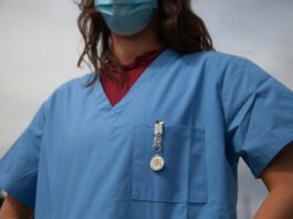 Une alliance appelle à la manifestation nationale des soins Vue rapprochée du buste d’un-e soignant-e portant une blouse médicale bleue et un masque chirurgical. On aperçoit un badge « Medical Alert » accroché à la poche poitrine et, sous la blouse, un haut bordeaux à col montant.
