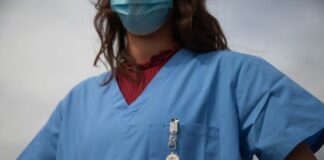 Une alliance appelle à la manifestation nationale des soins Vue rapprochée du buste d’un-e soignant-e portant une blouse médicale bleue et un masque chirurgical. On aperçoit un badge « Medical Alert » accroché à la poche poitrine et, sous la blouse, un haut bordeaux à col montant.