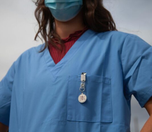 Une alliance appelle à la manifestation nationale des soins Vue rapprochée du buste d’un-e soignant-e portant une blouse médicale bleue et un masque chirurgical. On aperçoit un badge « Medical Alert » accroché à la poche poitrine et, sous la blouse, un haut bordeaux à col montant.