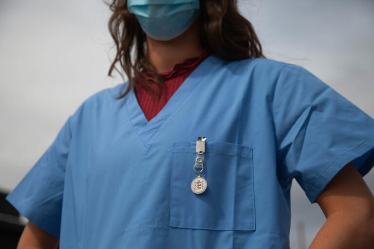 Vue rapprochée du buste d’un-e soignant-e portant une blouse médicale bleue et un masque chirurgical. On aperçoit un badge « Medical Alert » accroché à la poche poitrine et, sous la blouse, un haut bordeaux à col montant.
