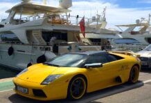 Les ultra-riches et le climat : ceux qui le détruisent doivent payer la facture Image montrant, au premier plan, une Lamborghini de sport décapotable jaune vif garée sur un quai, avec ses jantes noires à rebord jaune et sa plaque d’immatriculation espagnole. À l’arrière, un grand yacht de luxe blanc est amarré, sous un ciel bleu parsemé de nuages.