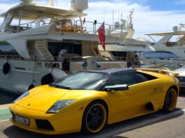 Les ultra-riches et le climat : ceux qui le détruisent doivent payer la facture Image montrant, au premier plan, une Lamborghini de sport décapotable jaune vif garée sur un quai, avec ses jantes noires à rebord jaune et sa plaque d’immatriculation espagnole. À l’arrière, un grand yacht de luxe blanc est amarré, sous un ciel bleu parsemé de nuages.
