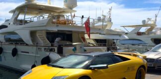 Image montrant, au premier plan, une Lamborghini de sport décapotable jaune vif garée sur un quai, avec ses jantes noires à rebord jaune et sa plaque d’immatriculation espagnole. À l’arrière, un grand yacht de luxe blanc est amarré, sous un ciel bleu parsemé de nuages.