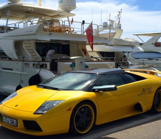 Les ultra-riches et le climat : ceux qui le détruisent doivent payer la facture Image montrant, au premier plan, une Lamborghini de sport décapotable jaune vif garée sur un quai, avec ses jantes noires à rebord jaune et sa plaque d’immatriculation espagnole. À l’arrière, un grand yacht de luxe blanc est amarré, sous un ciel bleu parsemé de nuages.