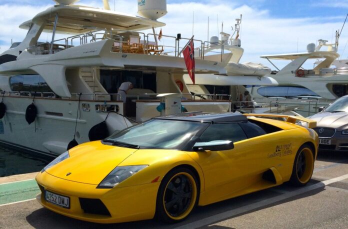 toni-pomar-mgf24cG_9sg-unsplash Image montrant, au premier plan, une Lamborghini de sport décapotable jaune vif garée sur un quai, avec ses jantes noires à rebord jaune et sa plaque d’immatriculation espagnole. À l’arrière, un grand yacht de luxe blanc est amarré, sous un ciel bleu parsemé de nuages.