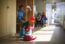 L’UDC veut démanteler le droit d’asile — les conséquences pour les réfugié-es sont dramatiques Dans un couloir lumineux d'un centre d'asile en Suisse alémanique au sol en bois, deux tout-petits sont assis par terre ; l’un d’eux lève les bras pour attraper un ballon rouge qui flotte au-dessus de sa tête. À gauche, une femme est assise sur un banc, les observant, et en arrière-plan deux autres adultes sont également assis, tournés vers les enfants.