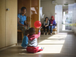 L’UDC veut démanteler le droit d’asile — les conséquences pour les réfugié-es sont dramatiques Dans un couloir lumineux d'un centre d'asile en Suisse alémanique au sol en bois, deux tout-petits sont assis par terre ; l’un d’eux lève les bras pour attraper un ballon rouge qui flotte au-dessus de sa tête. À gauche, une femme est assise sur un banc, les observant, et en arrière-plan deux autres adultes sont également assis, tournés vers les enfants.