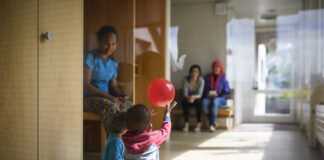 L’UDC veut démanteler le droit d’asile — les conséquences pour les réfugié-es sont dramatiques Dans un couloir lumineux d'un centre d'asile en Suisse alémanique au sol en bois, deux tout-petits sont assis par terre ; l’un d’eux lève les bras pour attraper un ballon rouge qui flotte au-dessus de sa tête. À gauche, une femme est assise sur un banc, les observant, et en arrière-plan deux autres adultes sont également assis, tournés vers les enfants.
